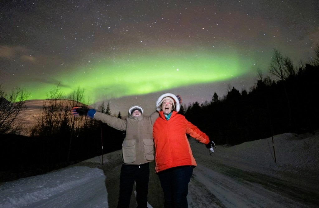 image of a woman and a teenage boy, lit up in a long-exposure photo of the northern lights. They are standing on a road near a forest with snow on the ground. 