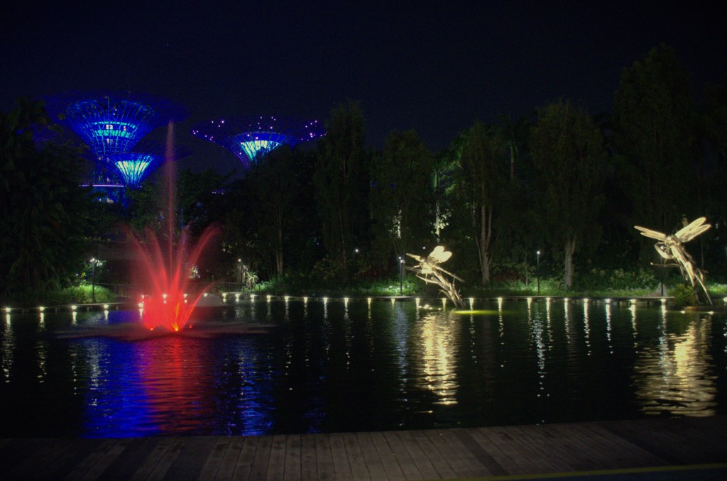 Image of Dragonfly Lake in Gardens by the Bay in Singapore. It is night and the lake has a red lit fountain with two gold dragonflies and the blue tree structures in the background