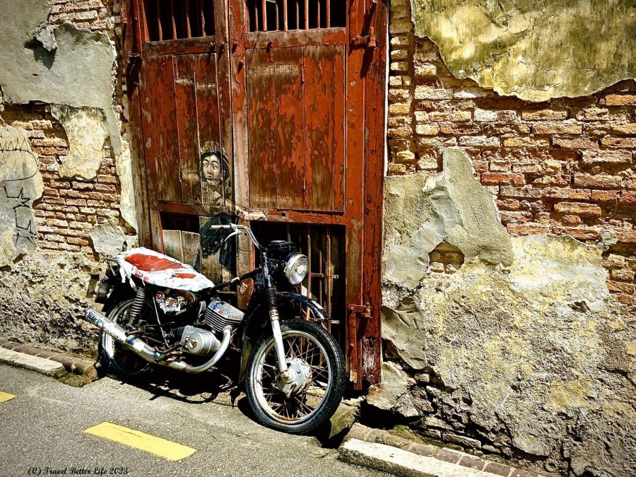 Photo of street art in George Town, Penang. Art includes painting of young boy with military-style bike helmet and real-life motorcycle leaning on wall at an angle to look like the painted child is atop the motorcyle.