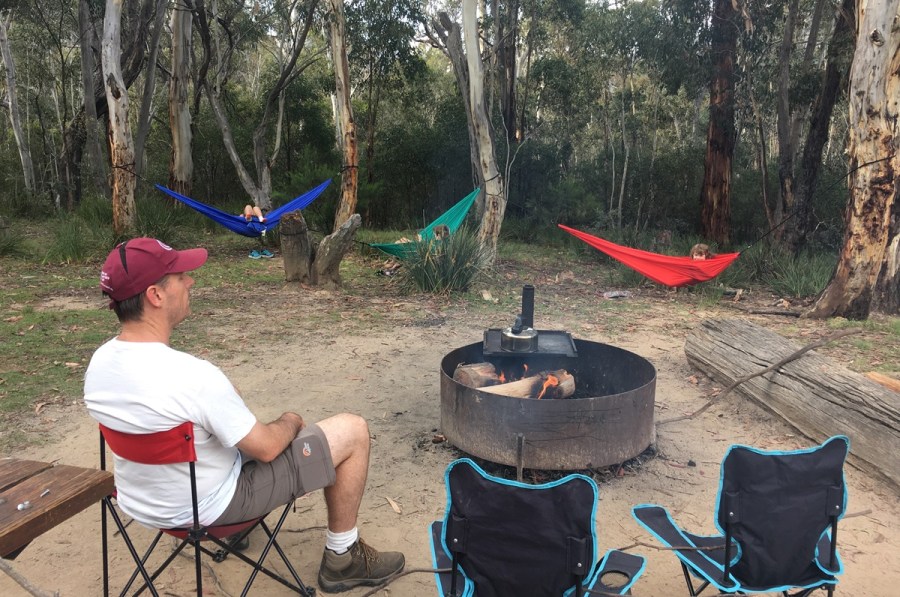 image of man sitting next to a firepit outdoors, with three hammocks tied to nearby trees. A billy kettle sits over the firepit, boiling water for tea. 