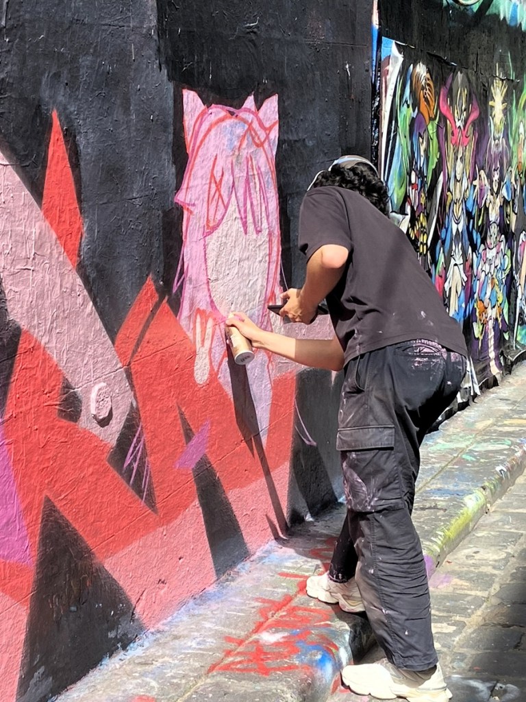 Photo of artist at work in Hosier Lane Melbourne spray painting a pink anime style kawaii girl with cat ears. Artist is known as @be3five