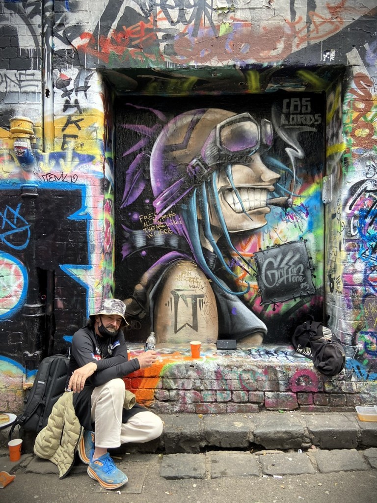 Photo of a man sitting in front of street art in Hosier Lane Melbourne. The street art shows a human head with a world war 2 style helmet and goggles smiling. The art is possibly by CBS Lord